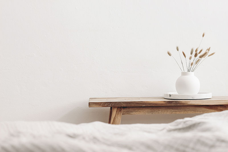 Learning Center 24 A minimalist scene with a wooden bench against a white wall, holding a round white vase filled with dried grass on a white tray. The foreground shows the edge of a soft, light-colored blanket.