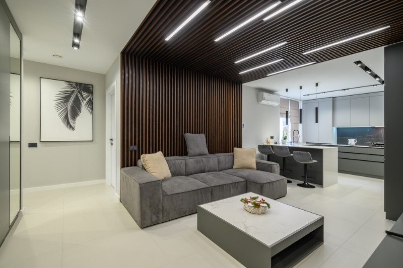 Modern living room with a gray sectional sofa, a low white coffee table, and a wall with vertical wooden slats. The open kitchen has bar stools and sleek cabinetry. Minimalist decor with neutral tones.