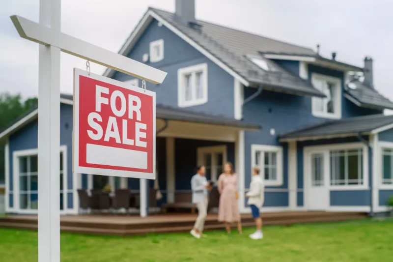 Bridge Home Loan 1 A red For Sale sign is displayed in front of a modern blue house. Three people are standing and talking on the porch in the background. The scene suggests a house showing or real estate transaction.